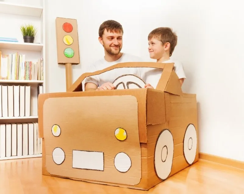 Một người lớn đang cùng trẻ em chơi với chiếc ô tô đồ chơi tự làm từ thùng giấy, thể hiện sự gắn kết gia đình qua đồ chơi DIY